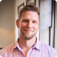 Smiling man with short light brown hair and beard wearing a pink and white checkered shirt, standing indoors near a window.