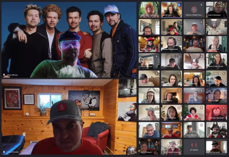 Screenshot of a virtual meeting with a group photo of five men on the top left, a man wearing a cap in a wooden room on the bottom left, and multiple participants in a video grid on the right.