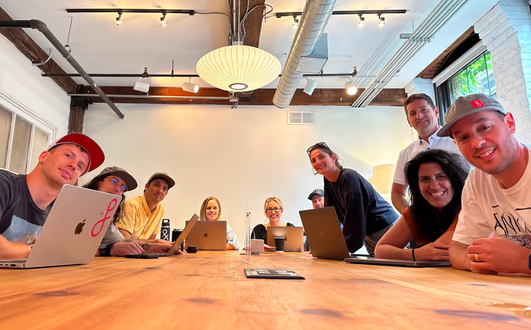 Group of nine people sitting and standing around a wooden conference table with laptops in a modern office space.