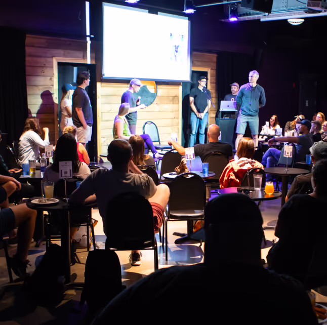 Group of people seated in a dimly lit room watching several presenters stand near a projection screen.