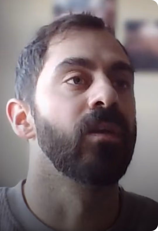 Close-up of a man with dark hair and a full beard looking slightly upwards, against a blurred indoor background with indistinct frames on the wall.
