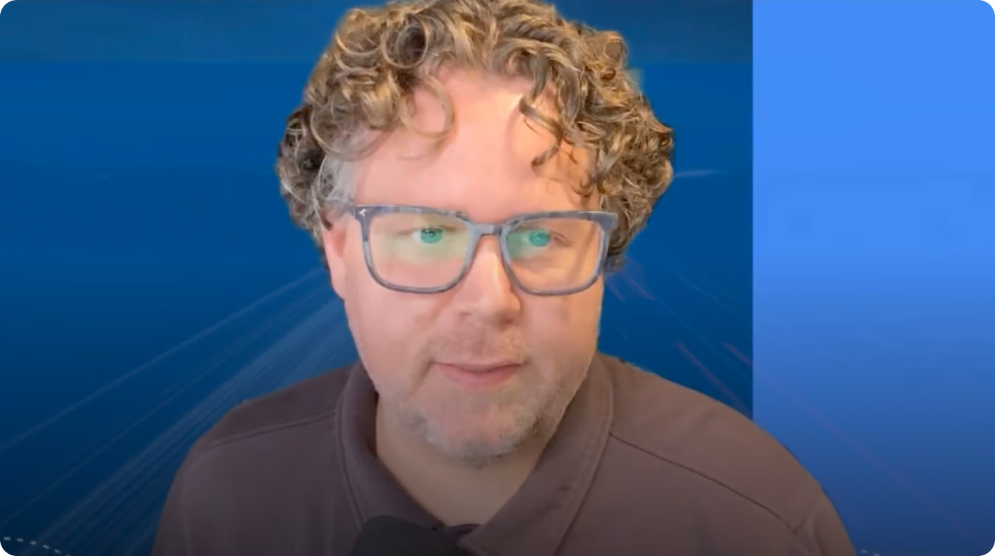 Man with curly hair and glasses wearing a brown shirt in front of a blue background.