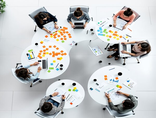 Sekiz kişinin etrafında oturduğu beyaz masalarda dizüstü bilgisayarlar, renkli yapışkan notlar ve grafikler var, toplantıda iş birliği yapıyorlar.