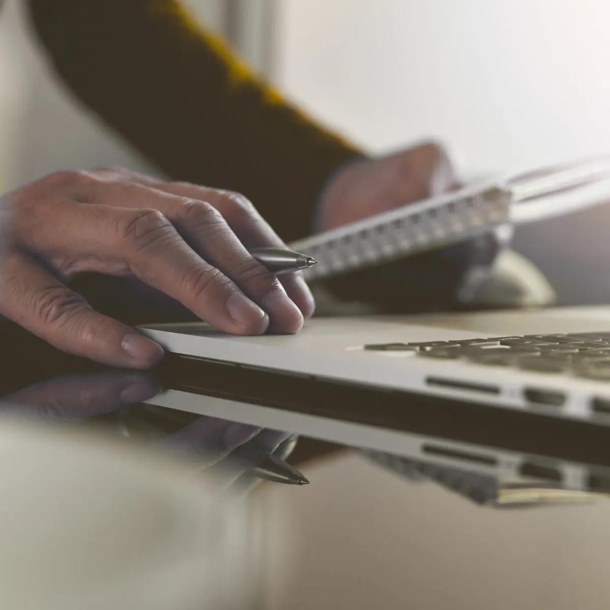 Hand mit Kugelschreiber auf Laptop-Tastatur, im Hintergrund Hand mit Notizblock verschwommen.