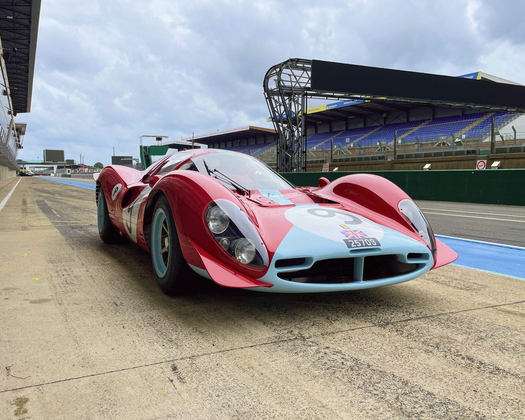 Ferrari de course classique rouge et bleu clair garée dans la voie des stands d'un circuit automobile sous un ciel nuageux.