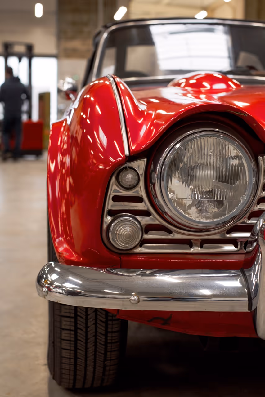 Gros plan sur l'avant d'une voiture ancienne rouge avec un phare rond et un pare-chocs chromé dans un garage.