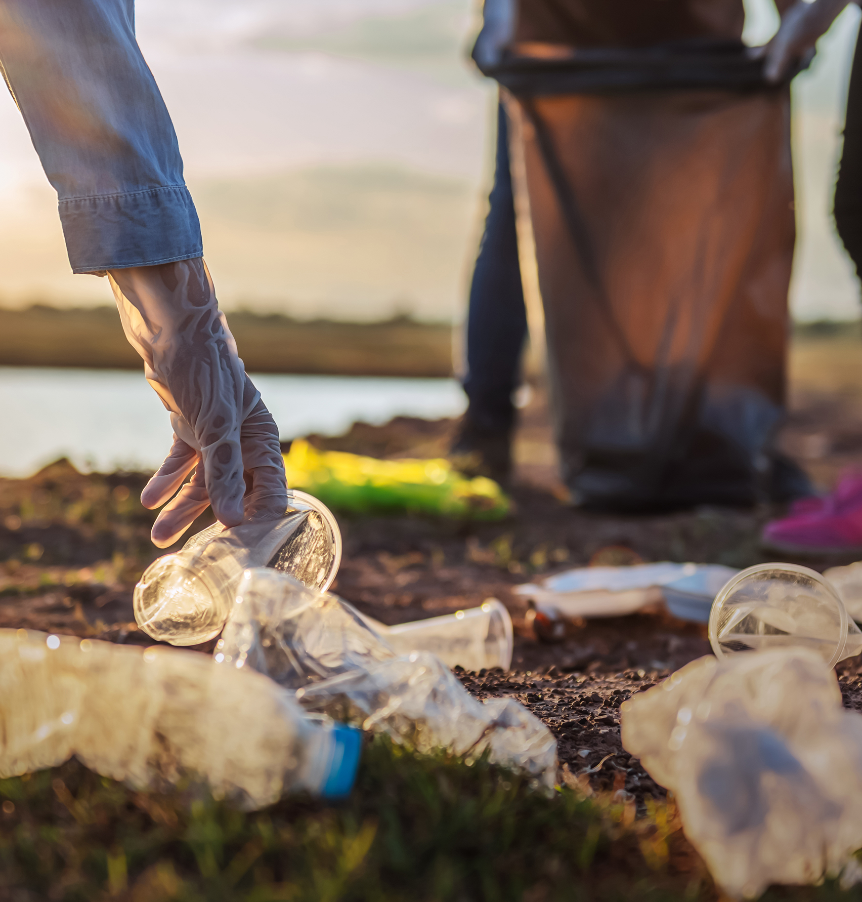 Person wearing a glove picking up plastic waste including bottles and cups during a cleanup near a body of water.