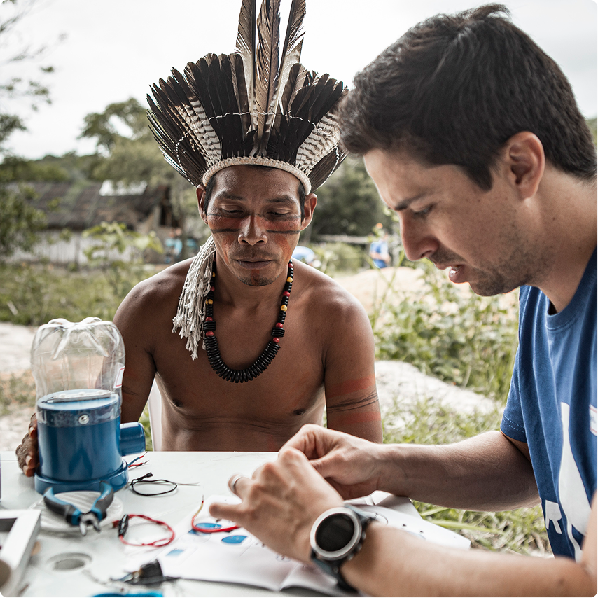 Indigenous man wearing a feathered headdress and traditional face paint sitting at a table outdoors, watching another man focused on assembling or fixing a device.