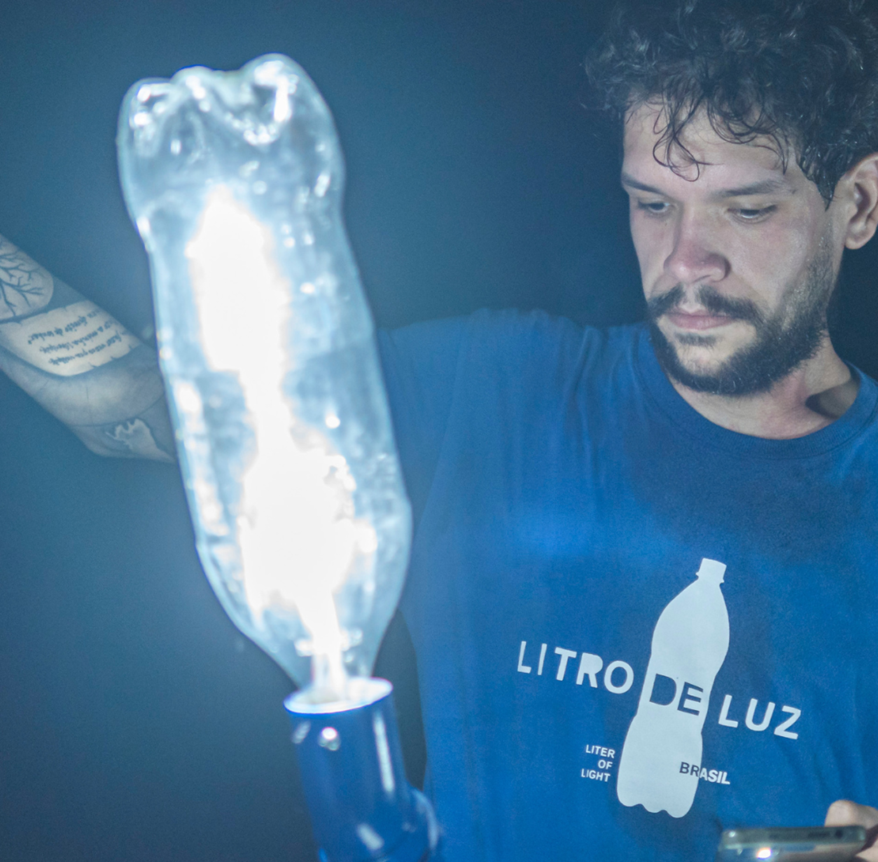 Man wearing a blue 'Litro de Luz' t-shirt holding a bright light source made from a plastic bottle.