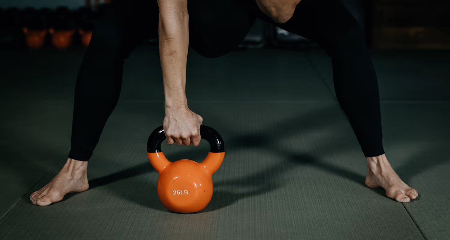 Person gripping an orange 25-pound kettlebell on a gym floor with bare feet wide apart.
