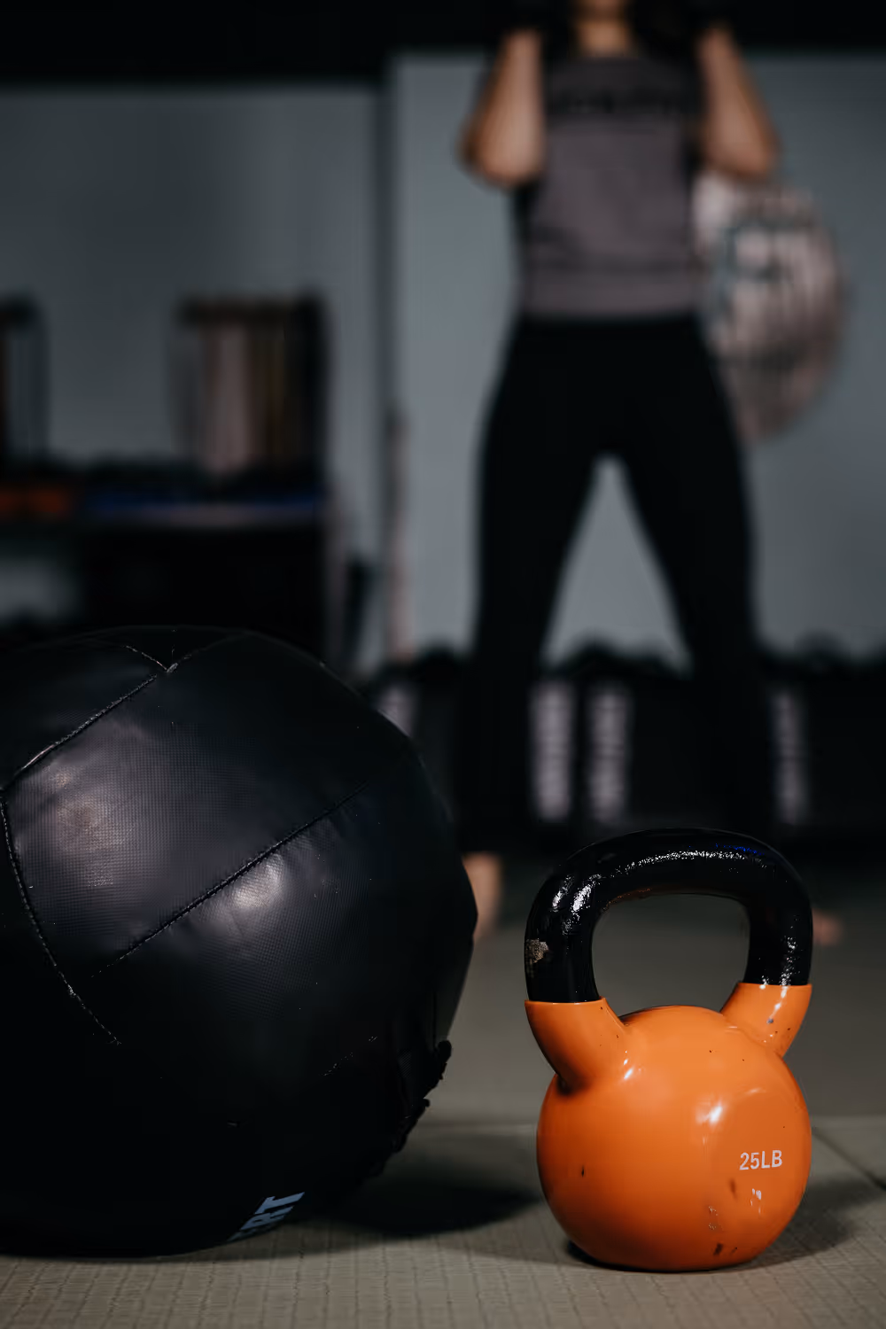 Orange 25-pound kettlebell and black medicine ball on gym floor, with a person lifting weights blurred in the background.