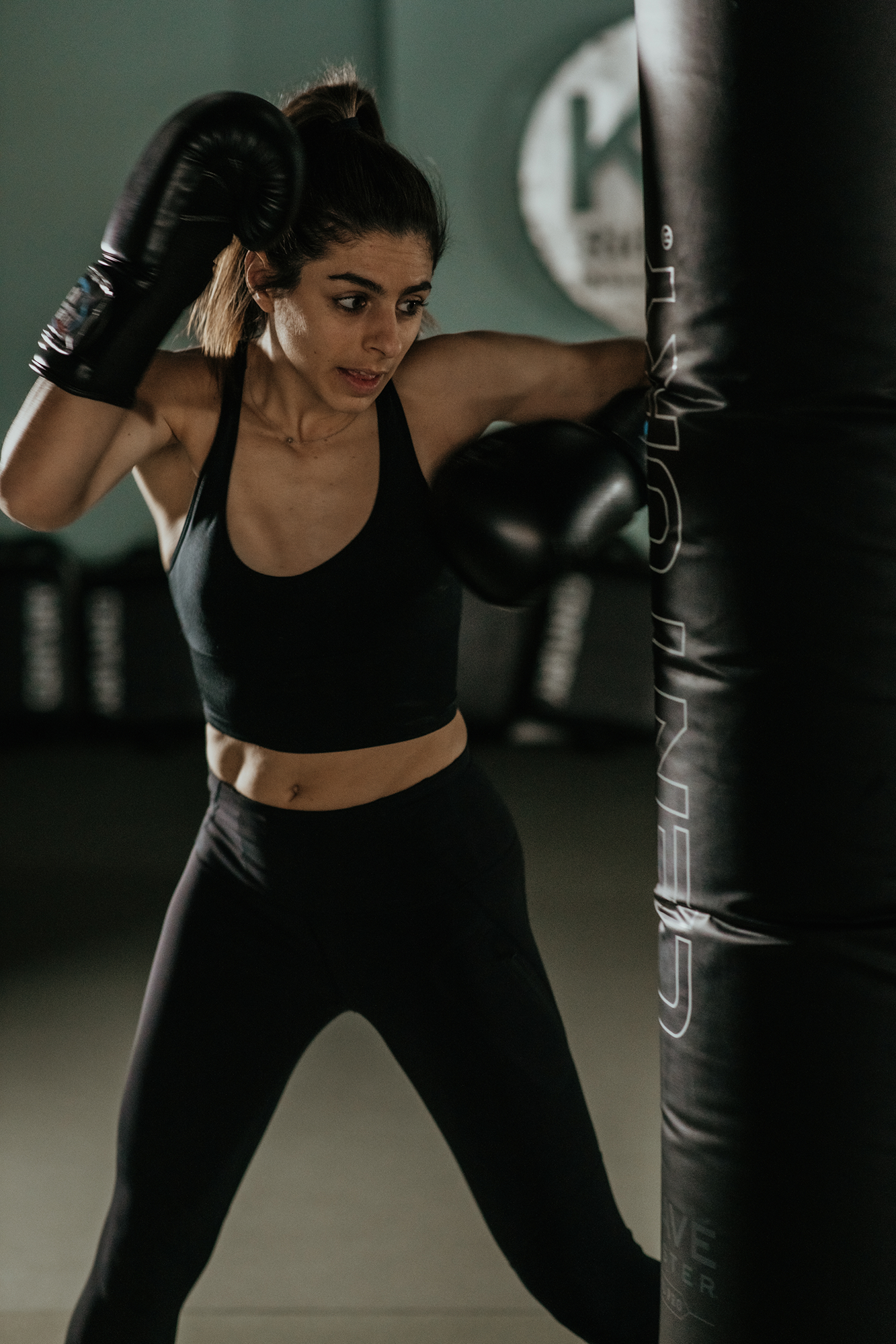 Woman in black sportswear and boxing gloves throwing a punch at a black punching bag in a gym.