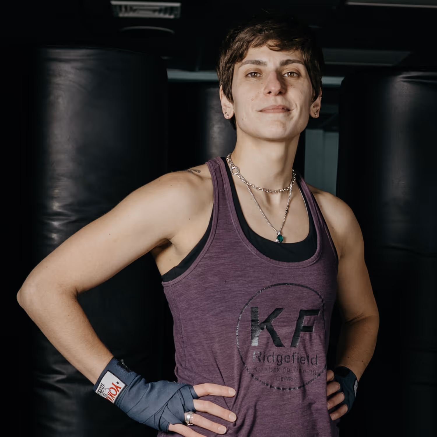 Fit person in purple tank top and hand wraps standing confidently in a gym with punching bags in the background.
