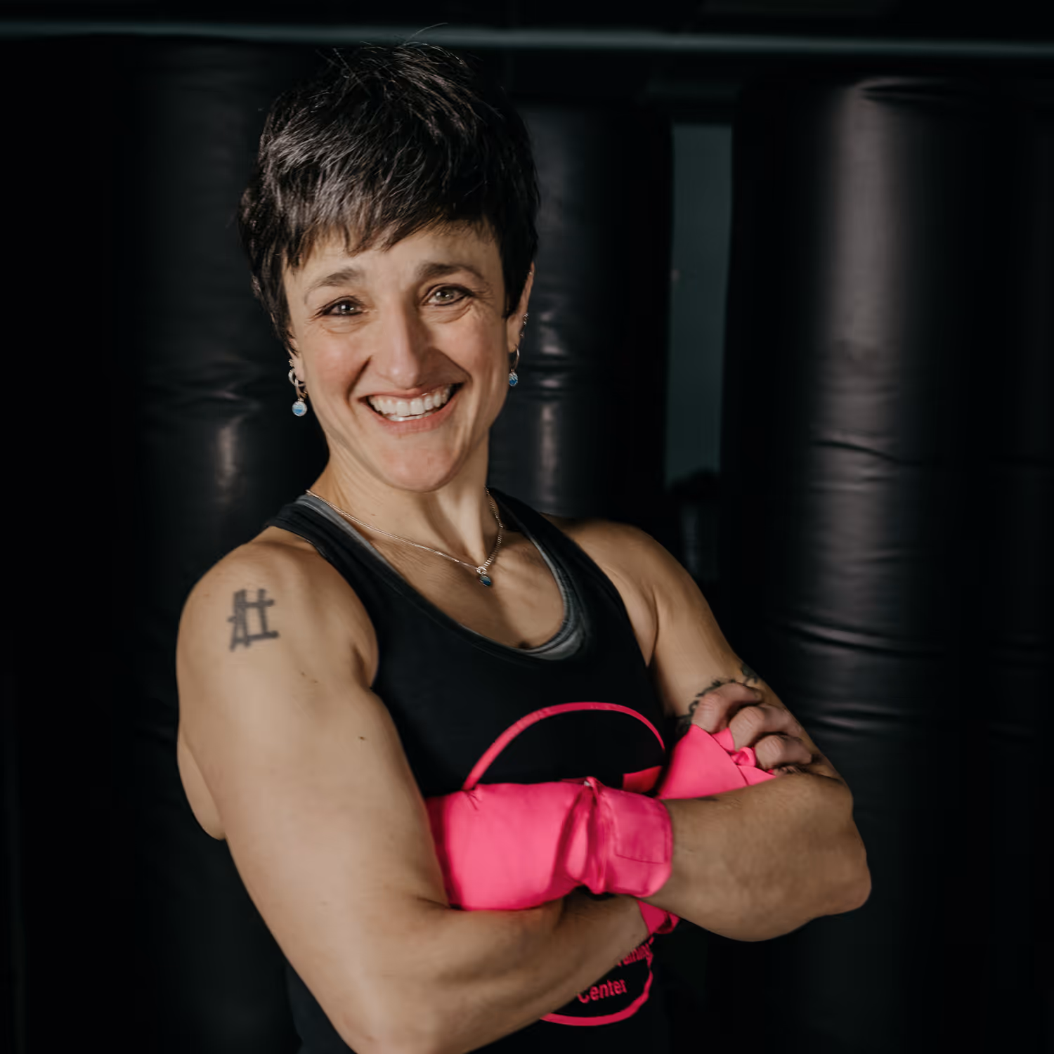 Smiling muscular woman in a black tank top wearing bright pink gloves with arms crossed in front of boxing bags.