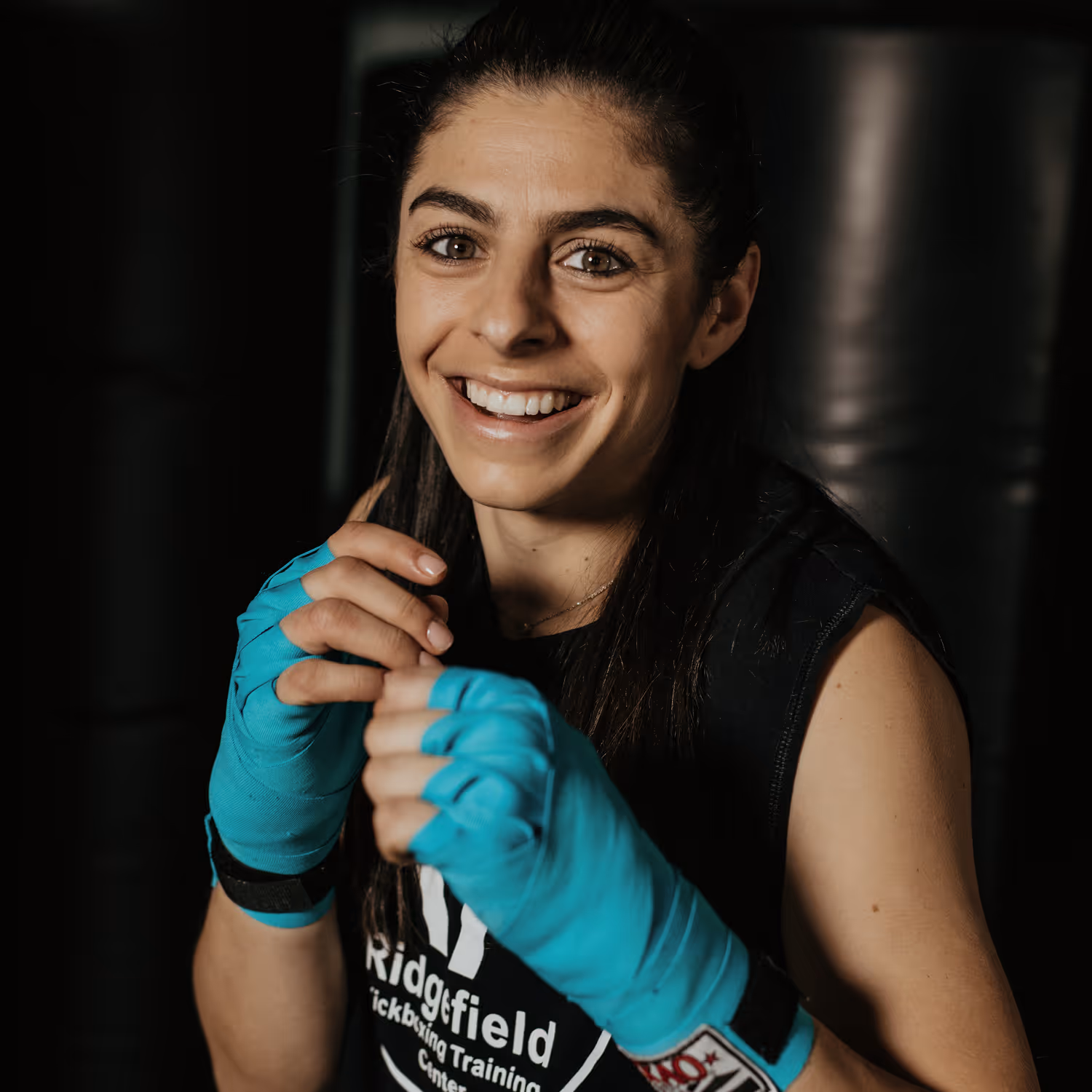 Smiling woman with long dark hair wearing turquoise hand wraps and a black sleeveless shirt preparing for boxing training.