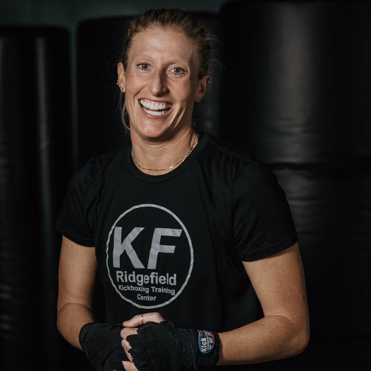 Smiling woman with wrapped hands wearing a black KF Ridgefield Kickboxing Training Center t-shirt.