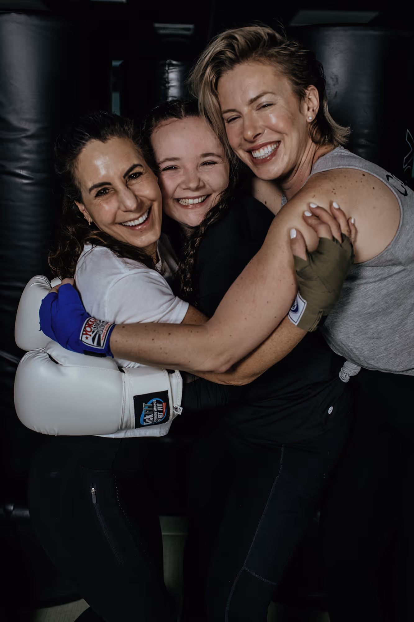 Three women wearing boxing gloves smiling and hugging each other inside a gym.