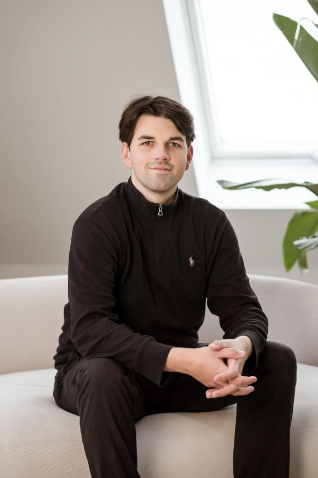 Rechtsanwalt Domenic Trybull in schwarzem Oberteil sitzt entspannt auf einem weißen Sofa vor einem Fenster mit Pflanzen im Hintergrund.