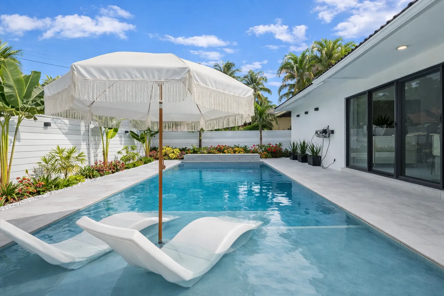A stylish outdoor patio area with a swimming pool, submerged sun loungers, and a large white fringed umbrella