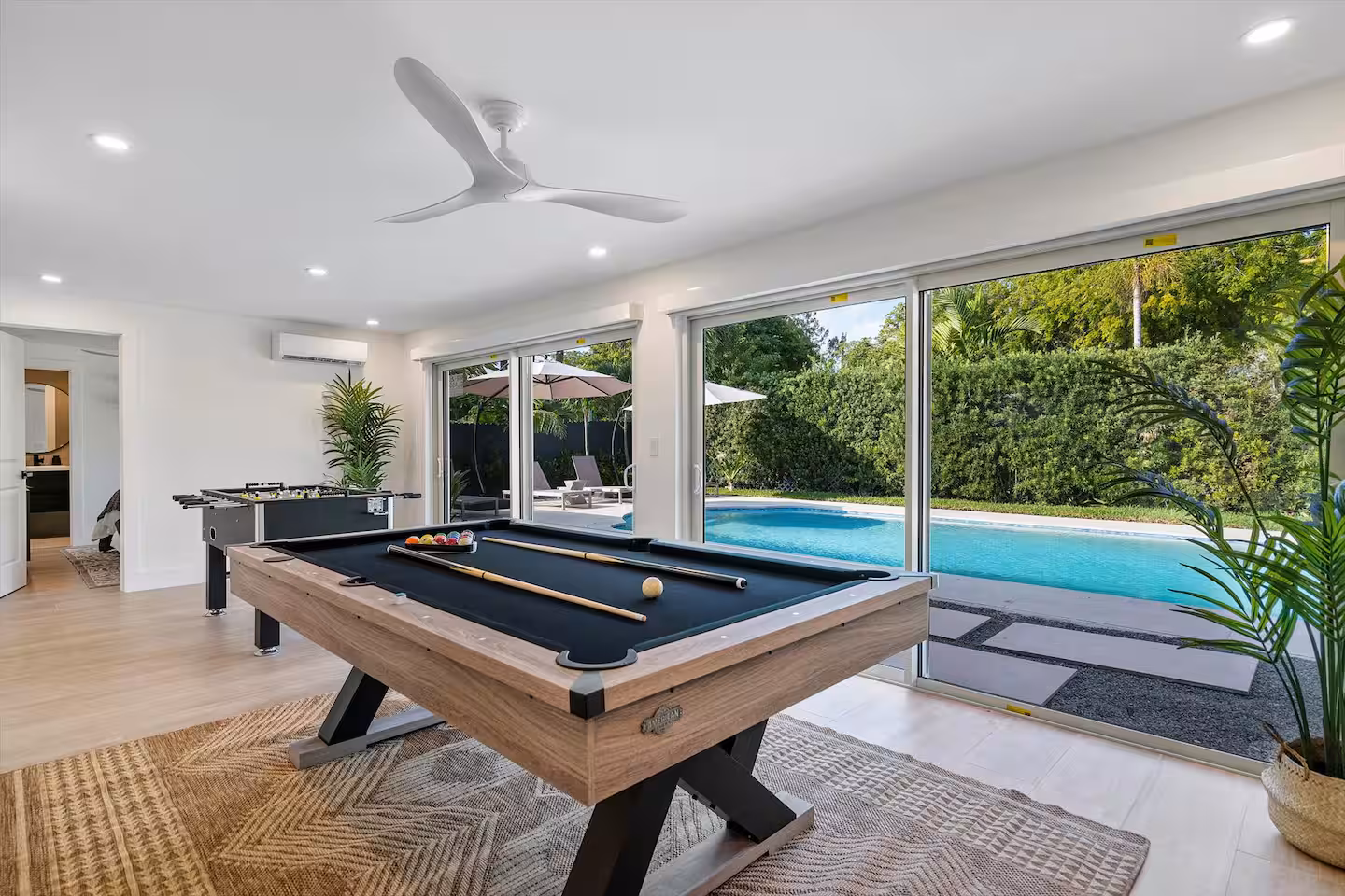 A modern indoor games room featuring a wooden billiard table and a foosball table, with large sliding glass doors overlooking a backyard swimming pool