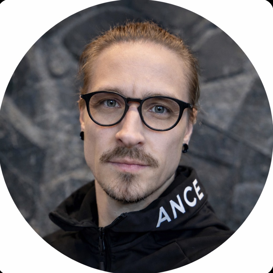 Mies, jolla on silmälasit ja ruskeat hiukset kiinni ponnarilla, musta takki, jossa näkyy kirjainyhdistelmä ANCE, vakava ilme.