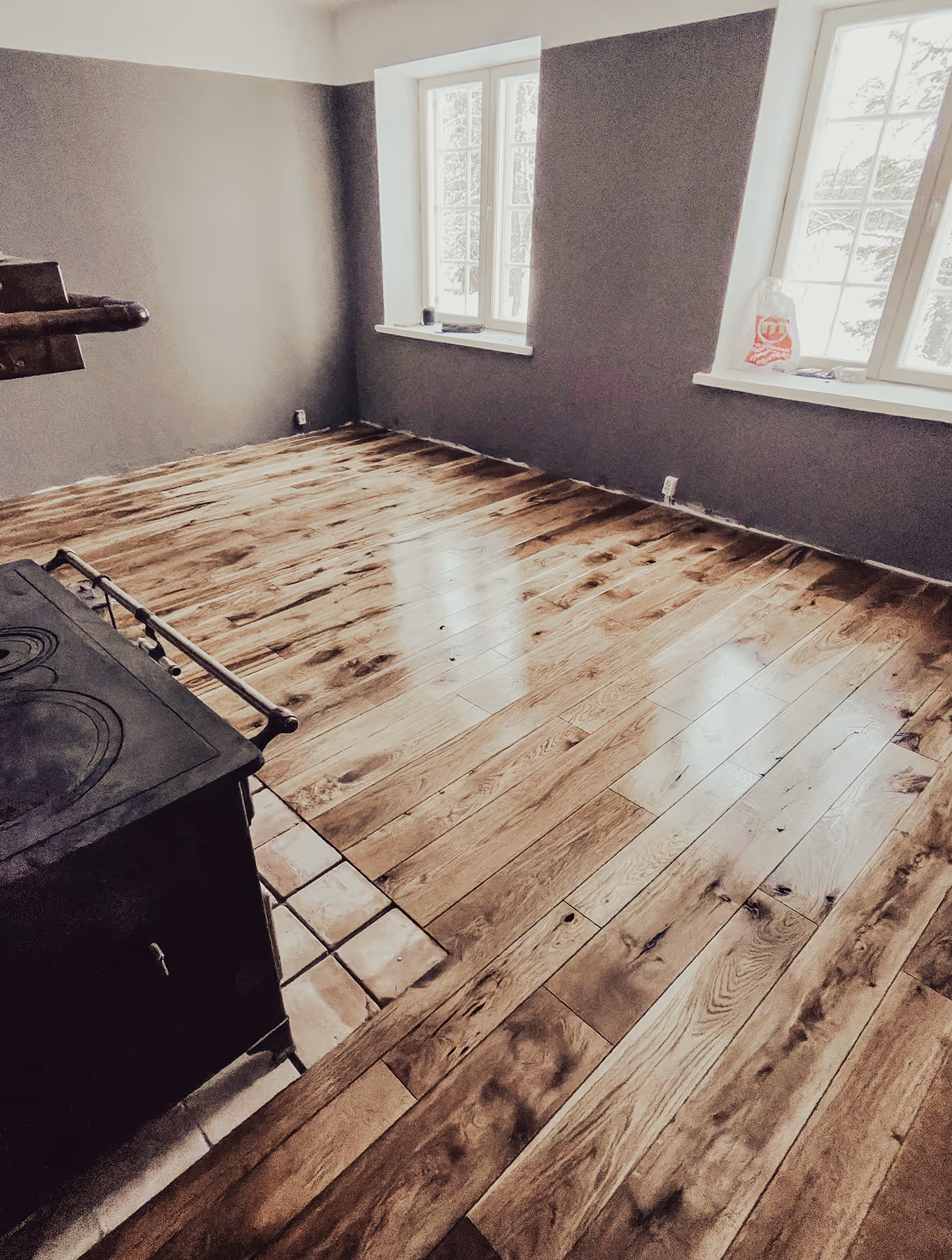 Tyhjä huone, jossa on uusi puulattia, kaksi suurta ikkunaa ja musta puuliesi vasemmassa reunassa.