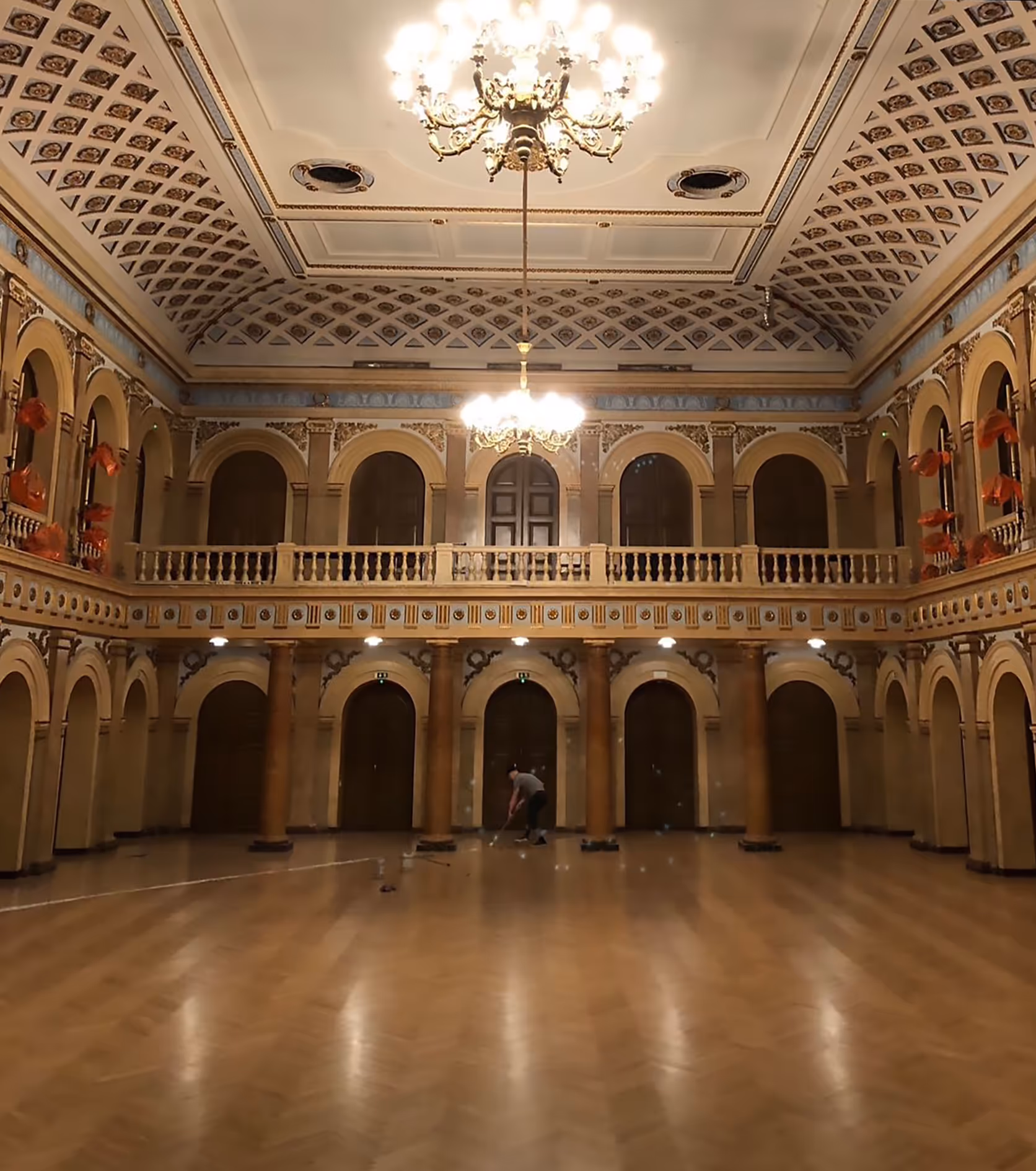 Suuri koristeellinen sali, jossa parvekkeet, pylväitä ja kattokruunut; henkilö lakaisee lattiaa.