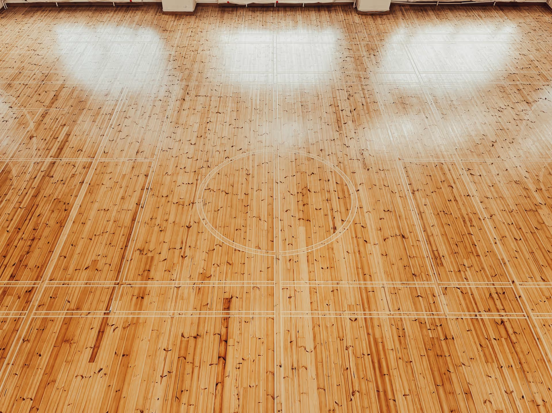 Tyhjä parkettilattia, jossa on koripallokentän rajaukset ja heijastuksia ikkunoista.