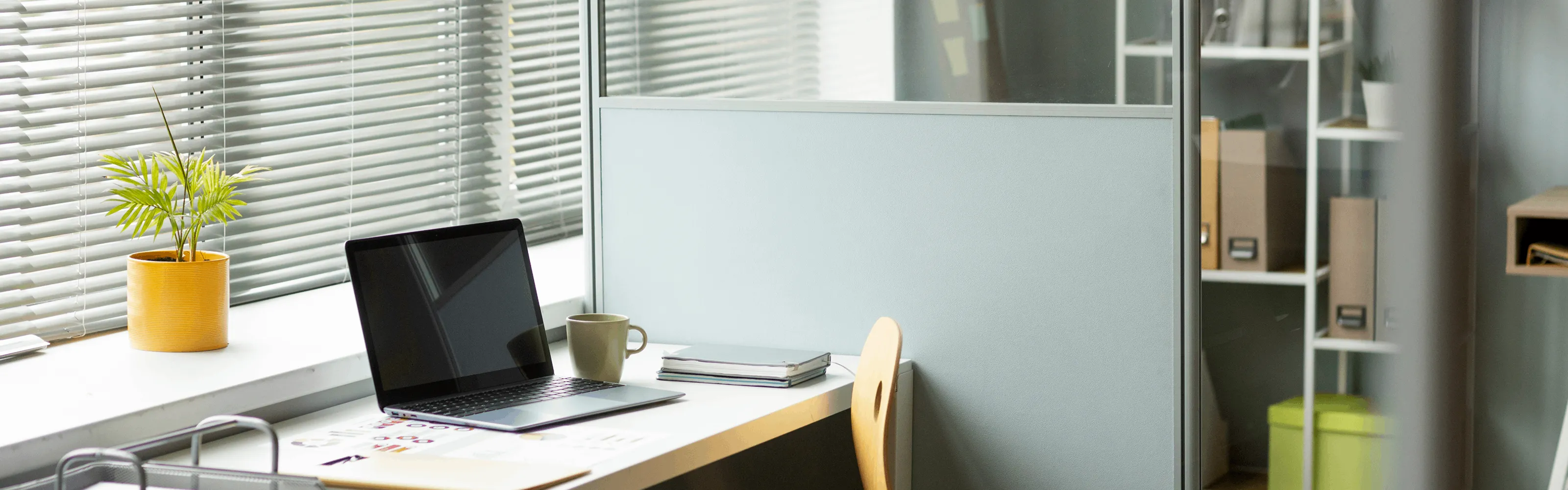 Modern office workspace with laptop, coffee mug, notebooks, and a potted plant by window blinds.