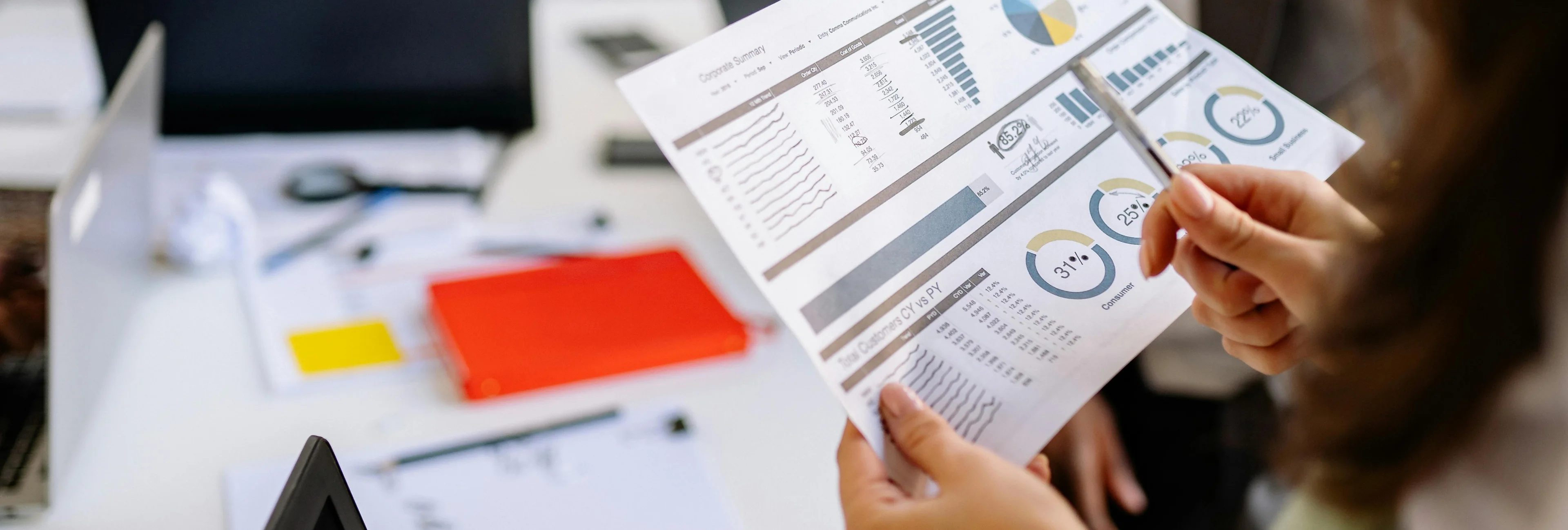 Person holding and pointing to a business report with charts and graphs on a desk with office supplies.