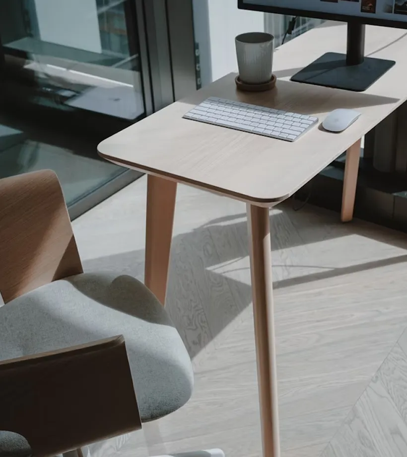 Wooden desk with a wireless keyboard, mouse, and a mug beside a computer monitor, next to a light gray cushioned chair.