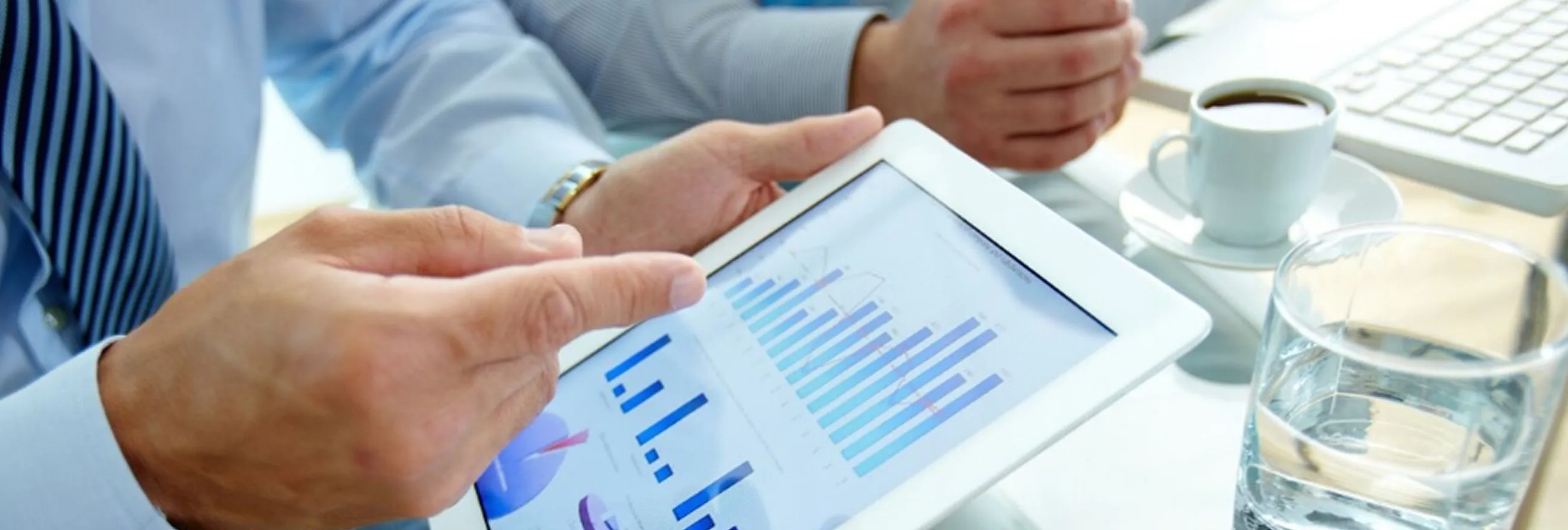 Two people in business attire reviewing charts and graphs on a tablet, with a cup of coffee and a glass of water nearby.