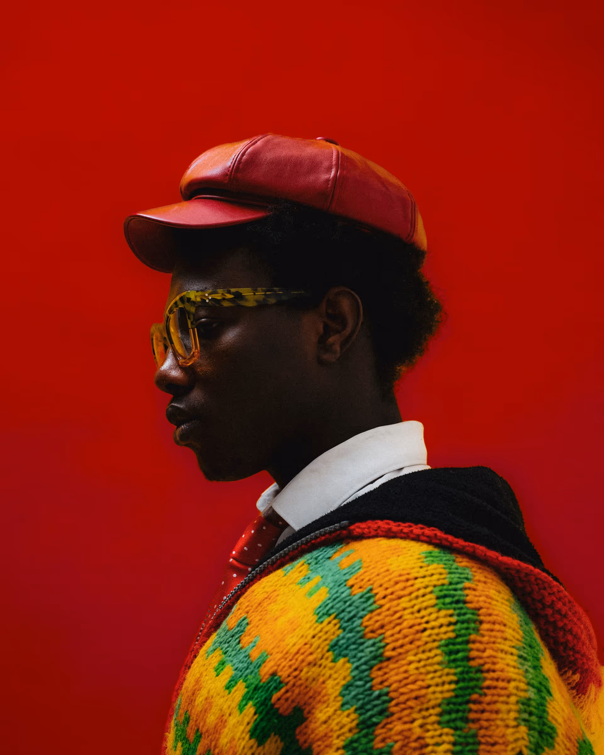 Side profile of a person wearing a red leather cap, yellow patterned glasses, white collared shirt, red tie, and a multicolored knitted sweater against a red background.