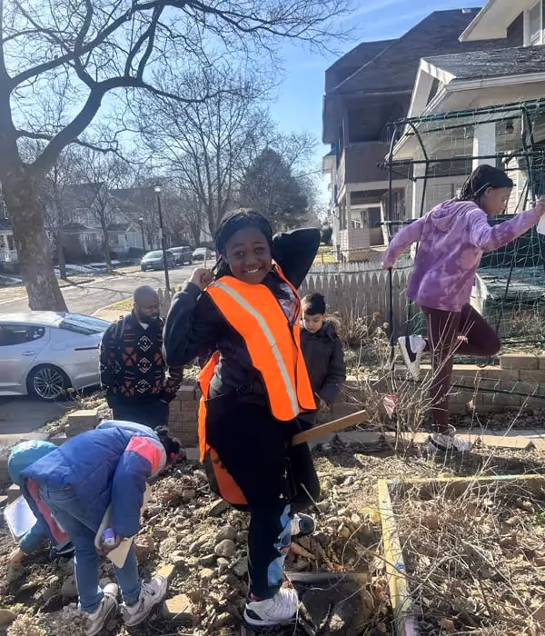 Children and an adult gardening outdoors on a sunny day, with one child smiling wearing an orange safety vest.