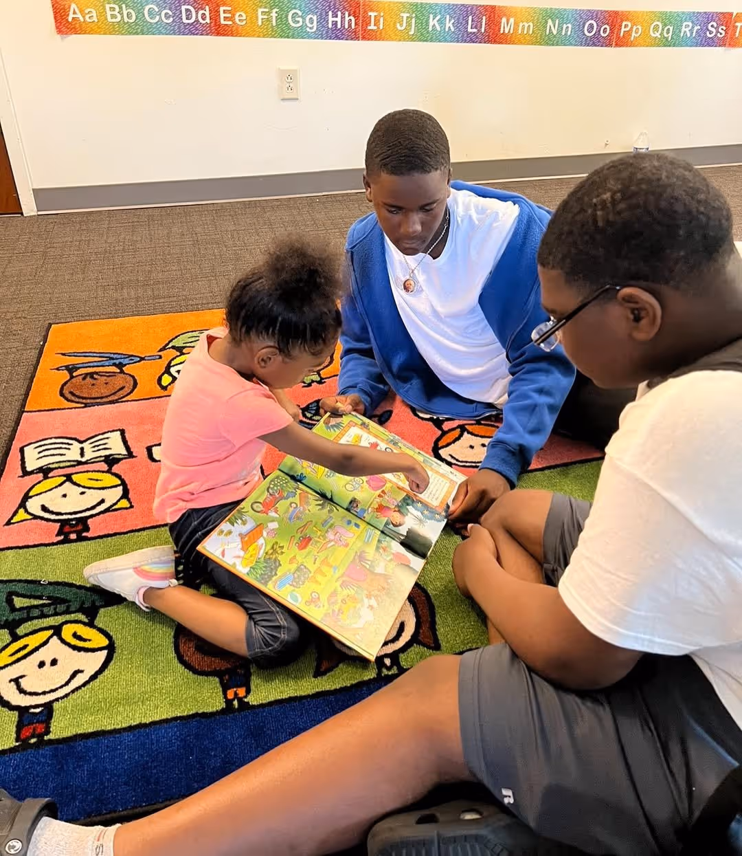 Three children sitting on a colorful alphabet rug reading and discussing a picture book together.