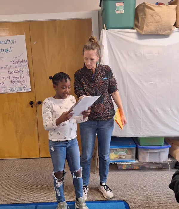 A young girl reads from a notebook while an adult woman stands beside her, looking at the notebook in a classroom setting.