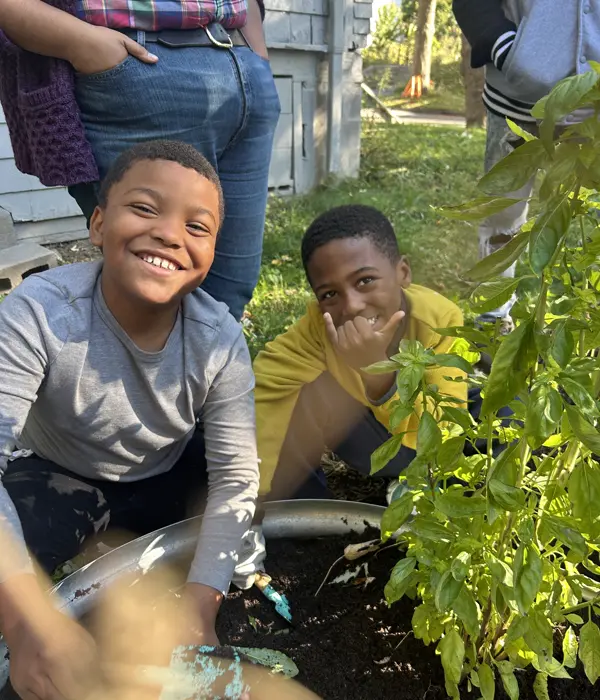 Two smiling boys gardening outdoors, one with hands in soil and the other making a hand gesture behind green plants.