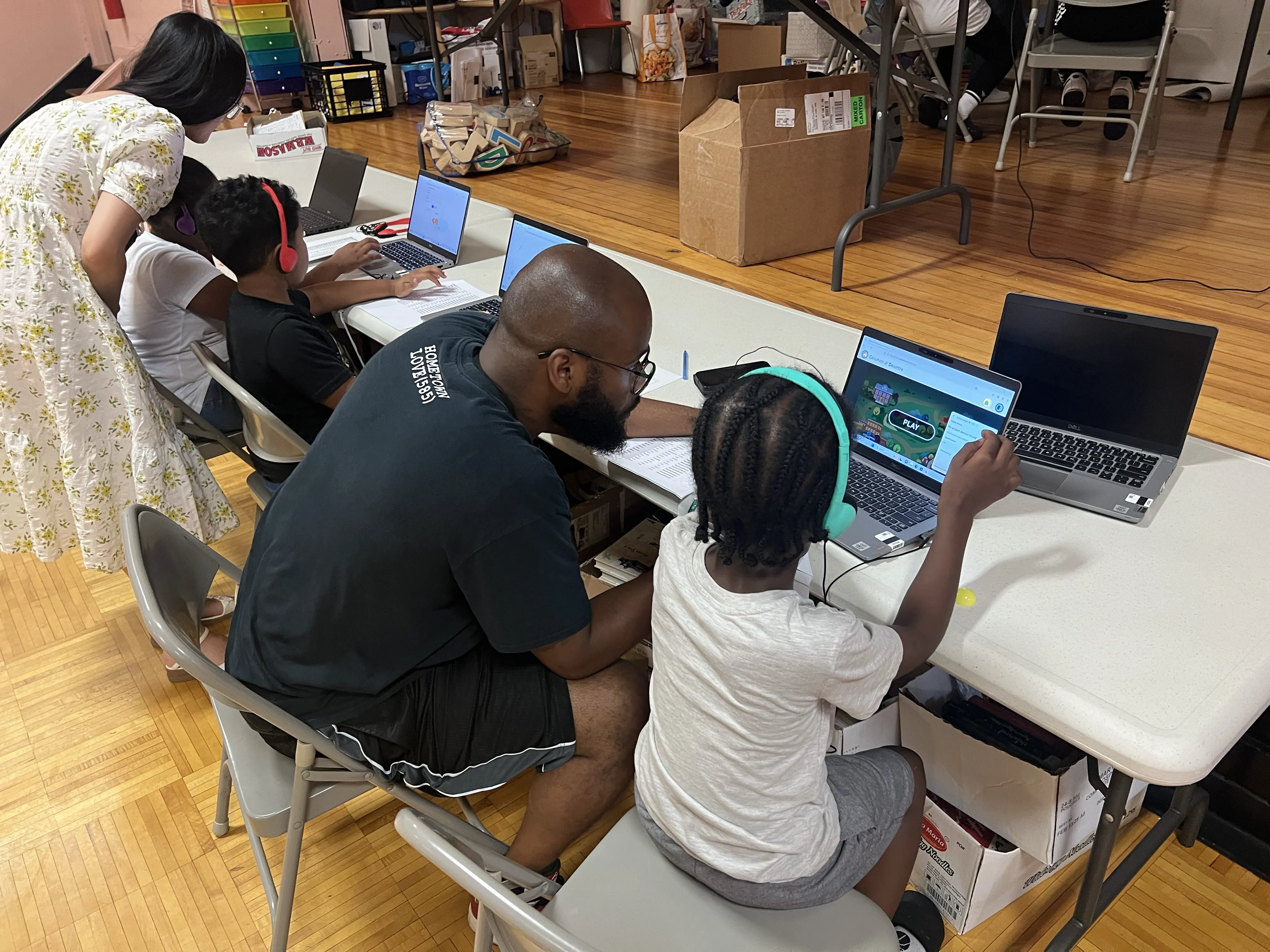A young girl reads from a notebook while an adult woman stands beside her, looking at the notebook in a classroom setting.