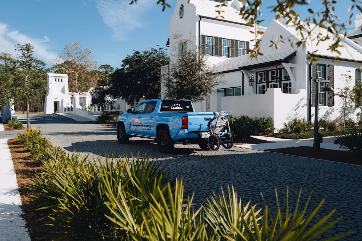 Blue Pool Captains pickup truck parked on a cobblestone road near white residential buildings with greenery in the foreground.
