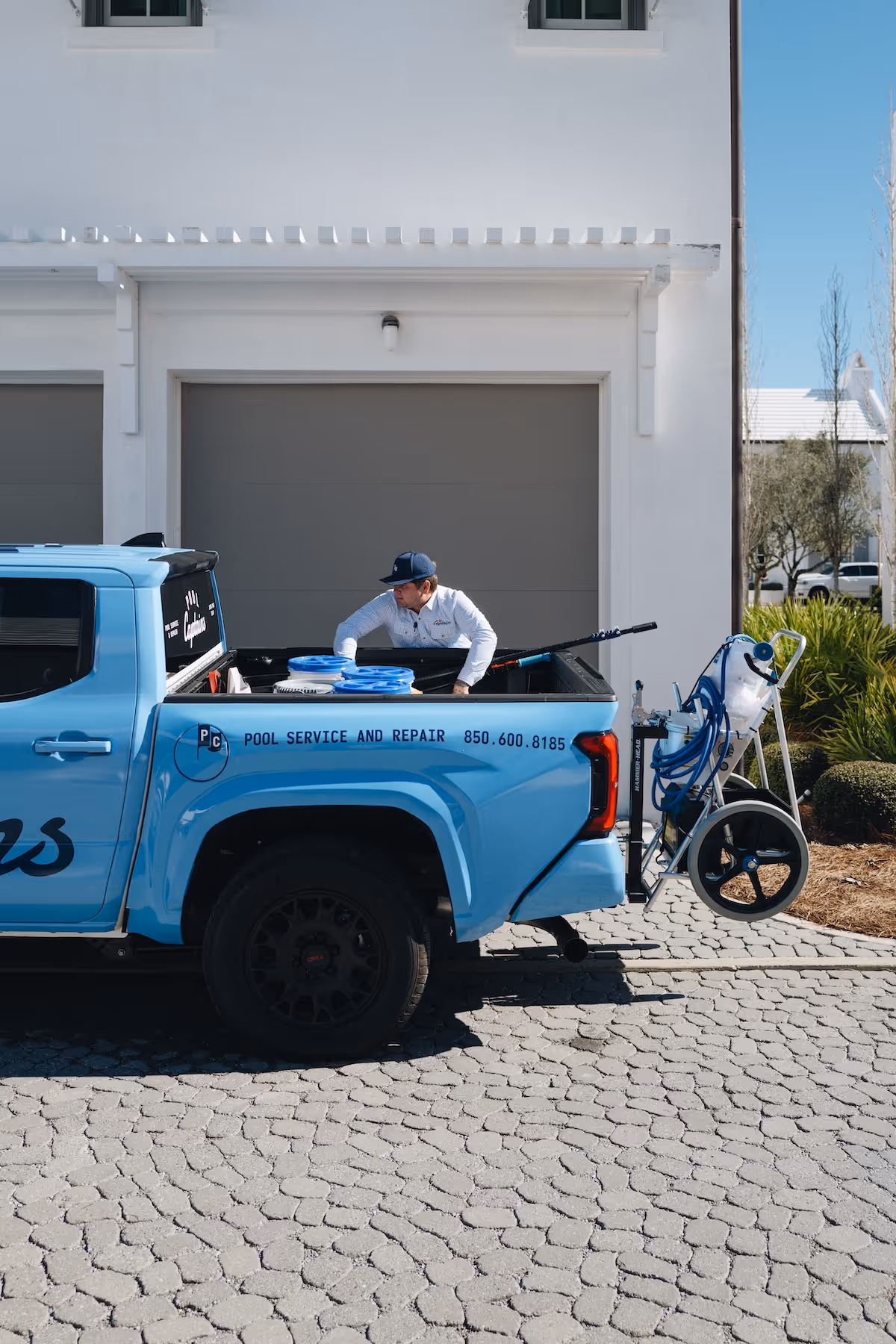 Technician loading equipment into the back of a blue pickup truck labeled for pool service and repair parked in front of a white garage.