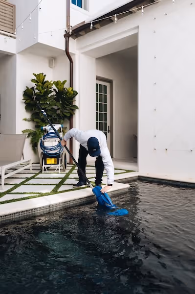 Person cleaning a pool with a blue net skimmer next to a modern white building patio.
