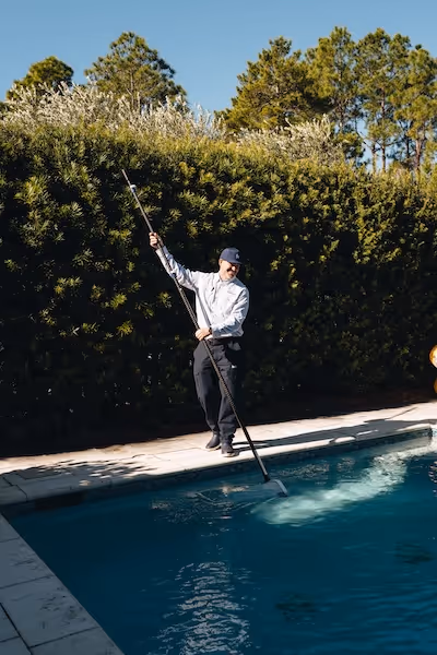 Man in uniform using a long pole to clean a swimming pool on a sunny day.