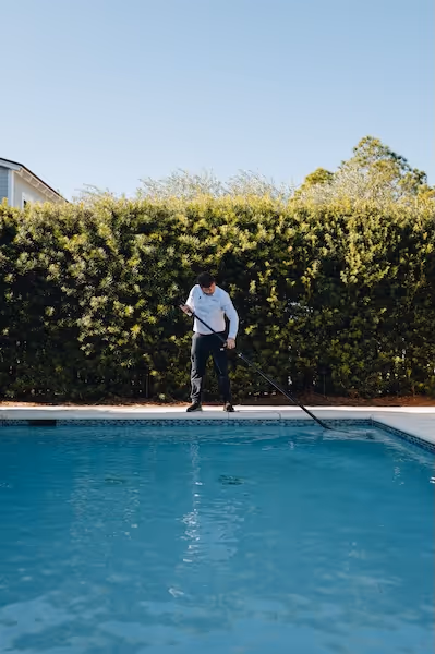 Pool captain cleaning a swimming pool with a long pole skimmer under clear blue sky.
