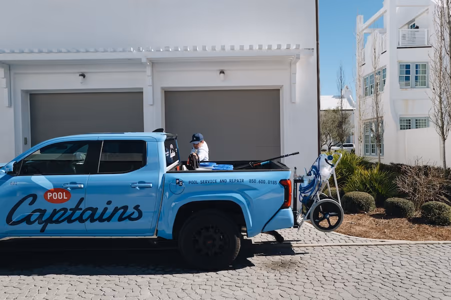 Blue Pool Captains pickup truck parked outside a white building with a person standing next to the truck bed with pool maintenance equipment attached to the back.