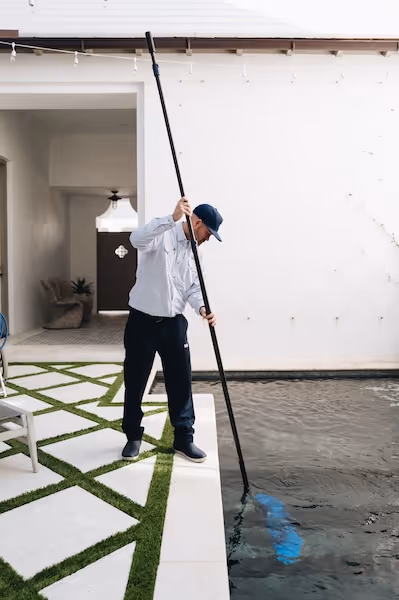Man in uniform cleaning a swimming pool with a long pole skimmer.