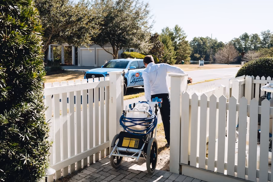 Man pushing pool maintenance cart through white picket gate with a blue Pool Captain truck parked in front of a suburban house.