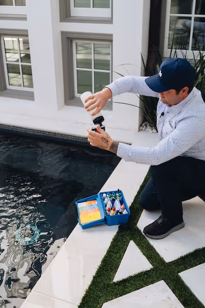 Man kneeling by a poolside testing pool water with a test kit containing various chemical bottles and color comparison cards.