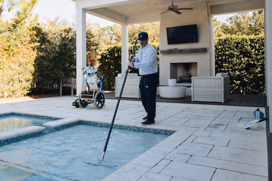A man in uniform using a long pole to clean a pool in a patio area.