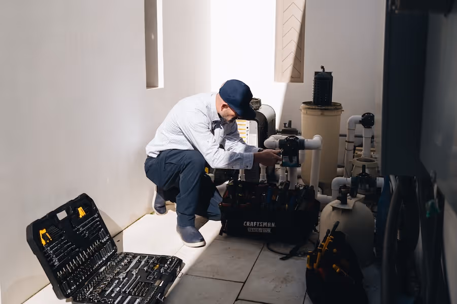 Technician kneeling and repairing plumbing pipes in a utility area with toolkits placed on the floor.