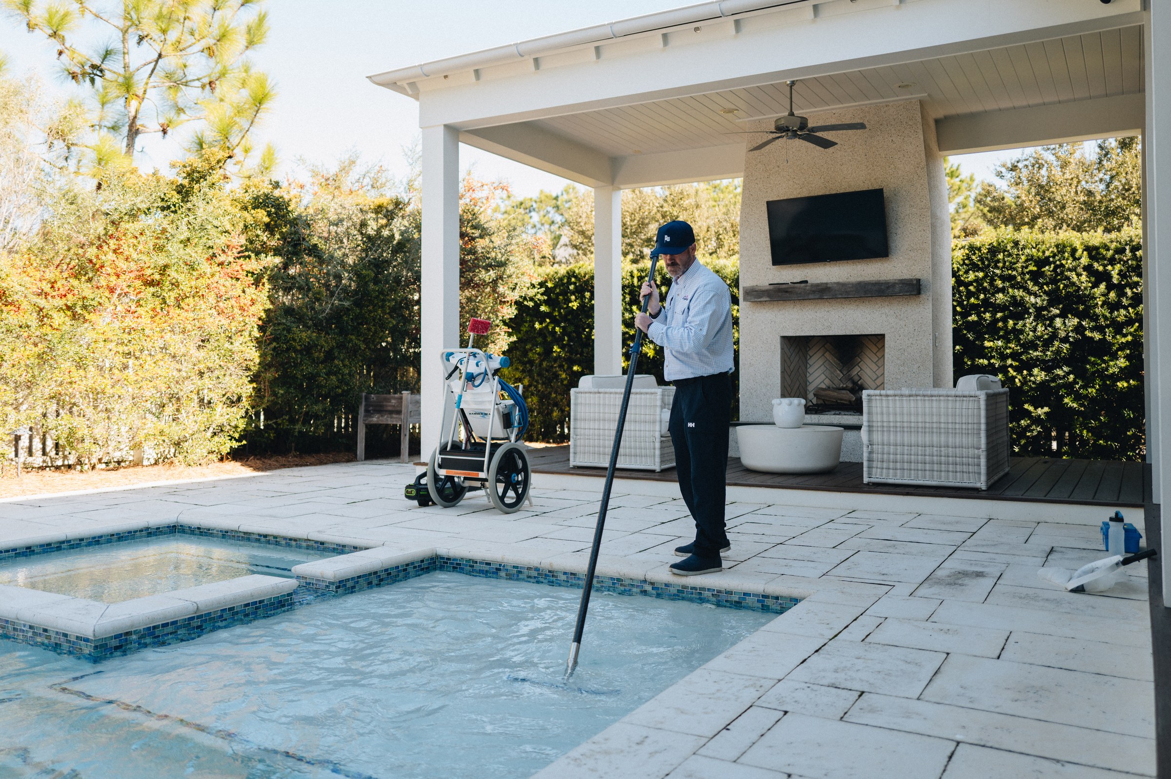 Wide view of a clear turquoise Emerald Coast swimming pool with spa corner as a Pool Captains technician brushes the water from the stone patio